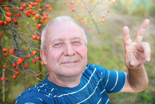 positive retired grandfather, makes selfie photo with a smartphone in the park. an elderly man walks in nature.