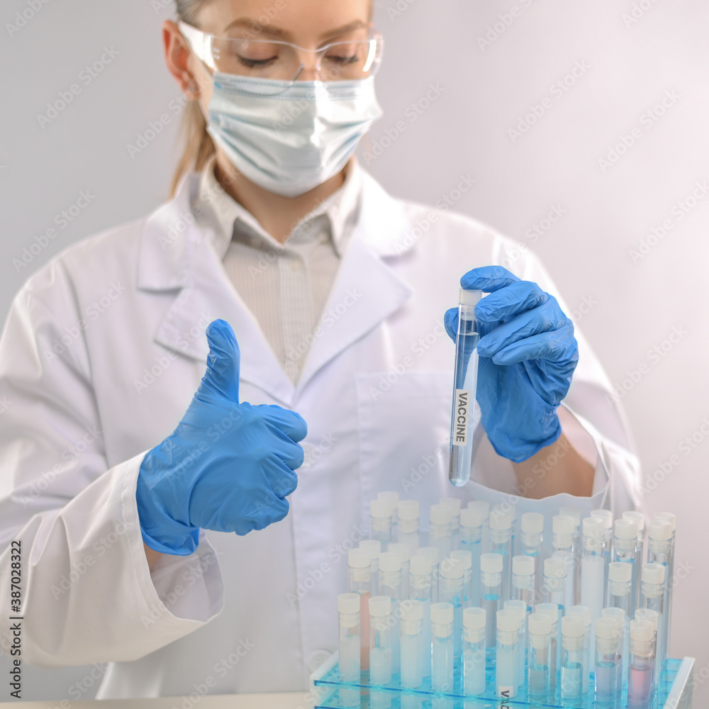 Close-up view of microbiologist in biohazard protective clothing keeps biological tubes with covid-19 vaccine and shows ok sign. Laboratory background. Healthcare and medical infection concept. Close-up view of microbiologist in biohazard protective clothing keeps biological tubes with covid-19 vaccine and shows ok sign. Laboratory background. Healthcare and medical infection concept.