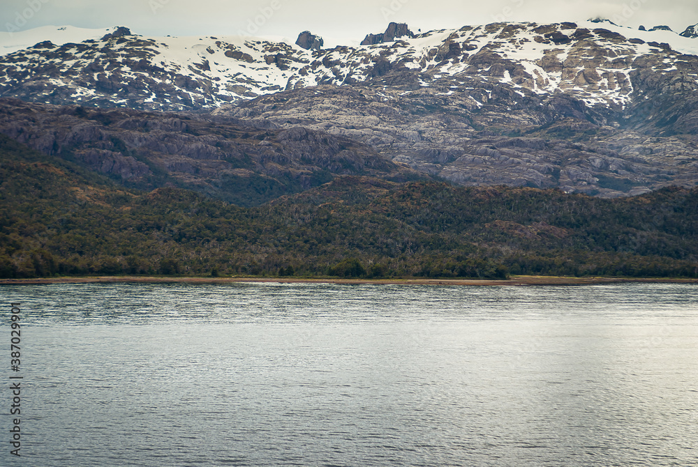 Sarmiento Channel, Chile - December 11, 2008: Amalia Glacier and Fjord. snow on range of black rock mountains behind belt of green foliage on hills and silver channel water.