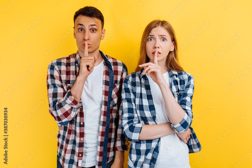 Young couple of friends, man and woman, holding index finger above lips, making silence gesture, on yellow background Young couple of friends, man and woman, holding index finger above lips, making silence gesture, on yellow background