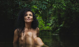 A young sexual naked woman with black curly hairs is sitting in the tropical creek surrounded by jungle. Image. Nude Portrait Photography. 