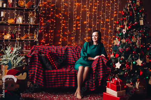 Full length of beautiful woman in elegant green dress on red checked sofa at decorated Christmas tree against lighting garland. Christmas time.