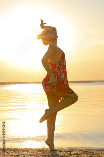 Beautiful girl stands on the beach  on one leg. She is near river  dressed into orange flower dress. It is summer sunset.