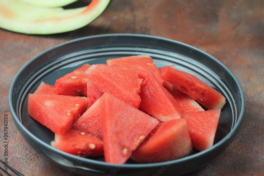 Buah Semangka or watermelon fruits, served on ceramic grey plate with fork beside, brown grainy background. Selective focus, close up. Buah Semangka or watermelon fruits, served on ceramic grey plate with fork beside, brown grainy background. Selective focus, close up.