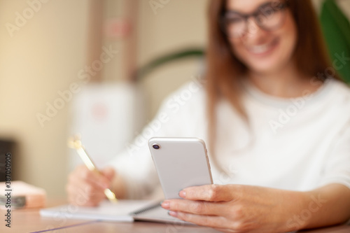 Concentrated young woman writing to do list in notepad and using task management mobile application