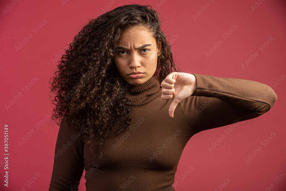 Portrait of a Brazilian woman with afro hairstyle and glamour makeup. Latin woman. Mixed race. Curly hair. Hair style. Red background. Thumbs down. Portrait of a Brazilian woman with afro hairstyle and glamour makeup. Latin woman. Mixed race. Curly hair. Hair style. Red background. Thumbs down.