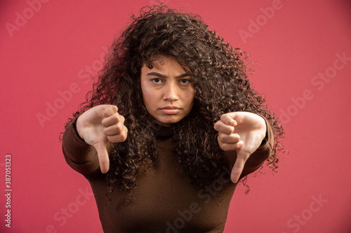 Portrait of a Brazilian woman with afro hairstyle and glamour makeup. Latin woman. Mixed race. Curly hair. Hair style. Red background. Thumbs down.