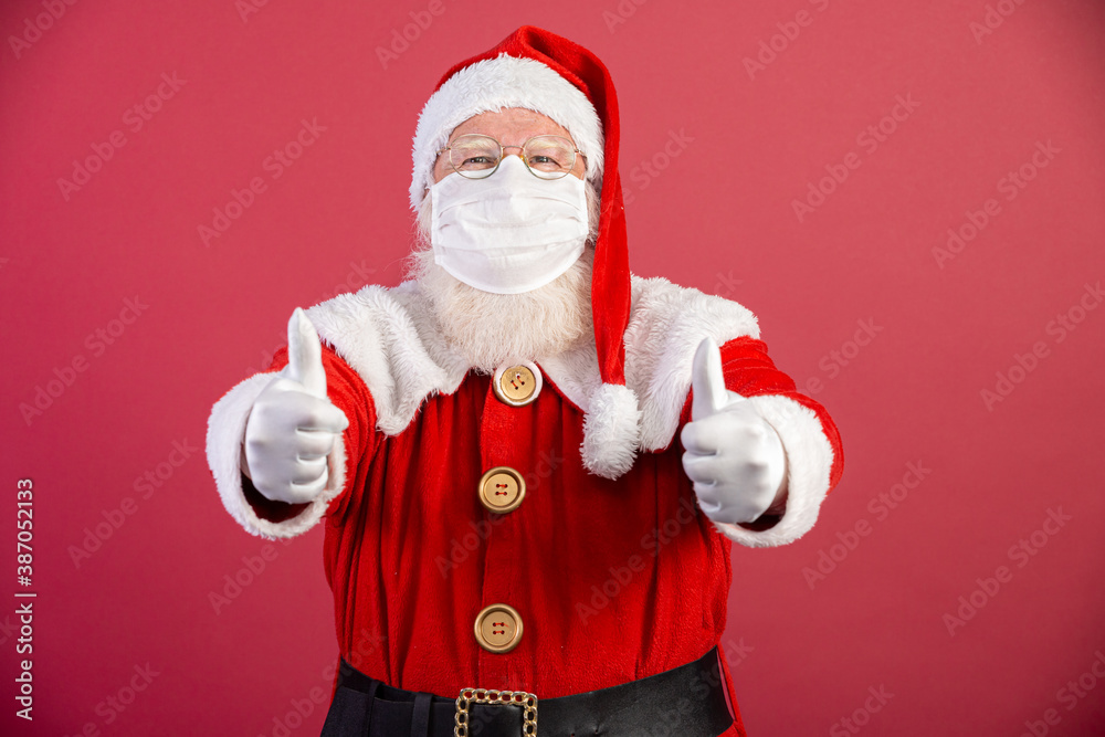 Real Santa Claus with red background, wearing protective mask, glasses and gloves with thumbs up. Christmas with social distance. Covid-19 - obrazy, fototapety, plakaty 