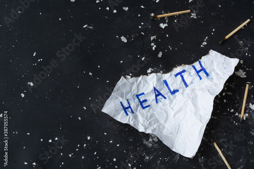 Crumpled burnt sheet of paper with inscription "Health" on black background with scattered ash and burnt matches . Health neglect and harm to health concept. Copy space. Top view