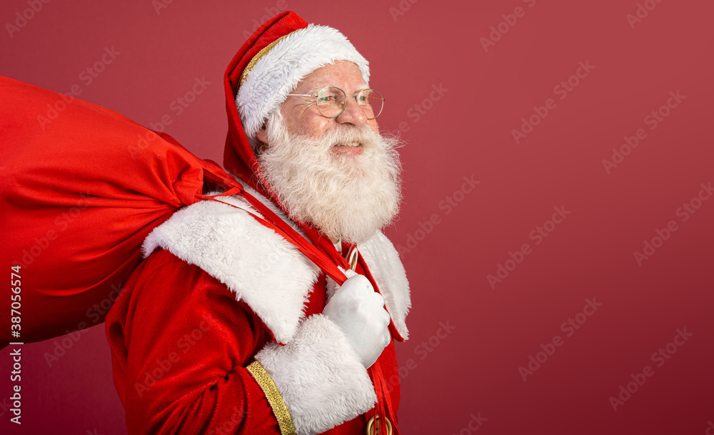Real Santa Claus with a red background, wearing glasses, gloves and a hat looking side.