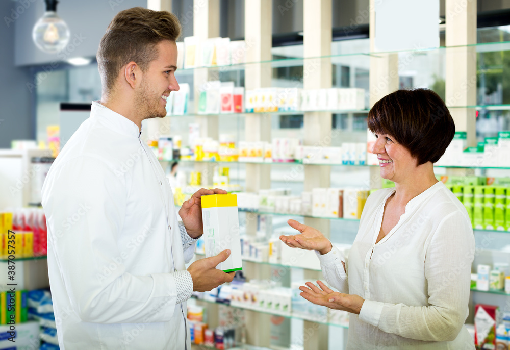 Happy adult man druggist in white coat giving advice to customers in pharmacy. Focus on the woman.