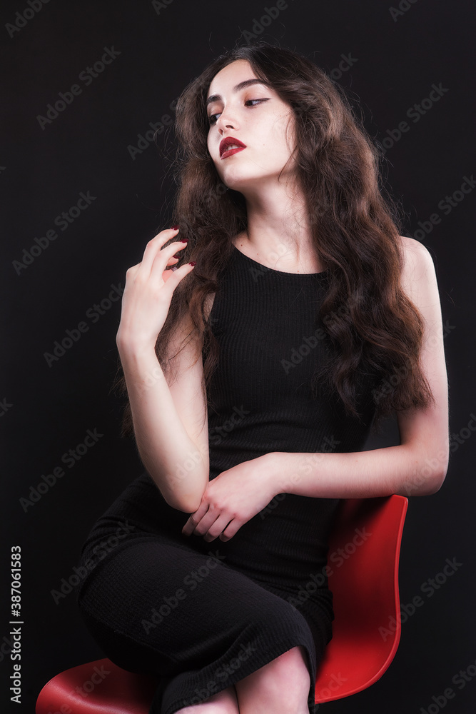 Beautiful slender young brunette with bright makeup in a black dress on a red chair Beautiful slender young brunette with bright makeup in a black dress on a red chair
