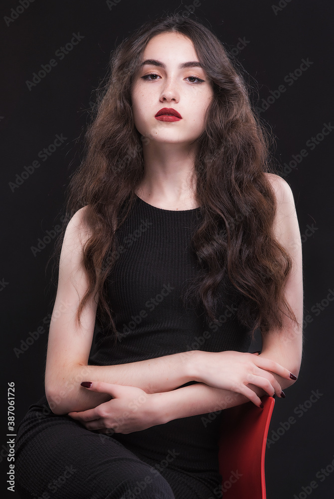 Fototapeta premium Beautiful slender young brunette with bright makeup in a black dress on a red chair