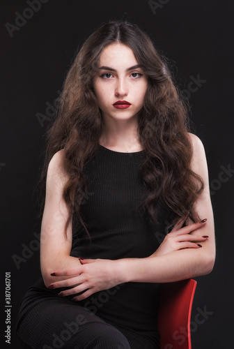 Beautiful slender young brunette with bright makeup in a black dress on a red chair