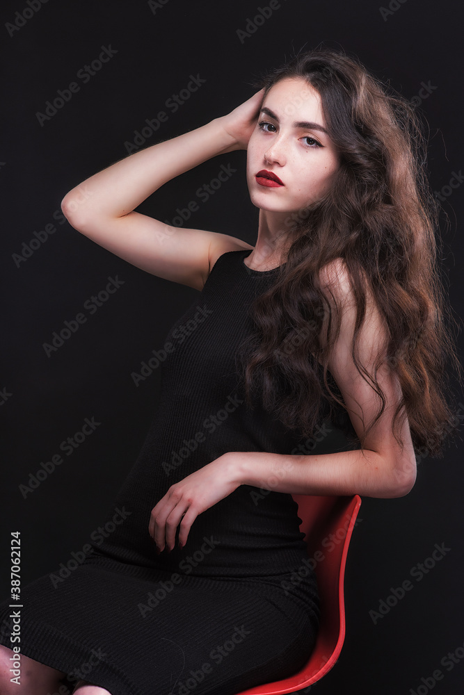 Beautiful slender young brunette with bright makeup in a black dress on a red chair