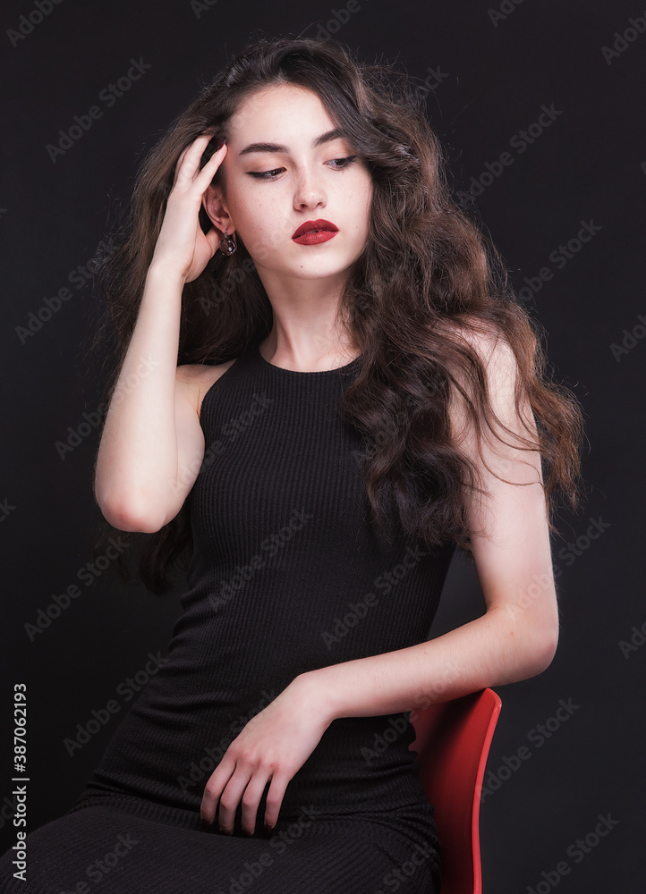 Fototapeta premium Beautiful slender young brunette with bright makeup in a black dress on a red chair