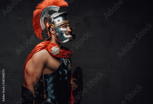 Obraz na plátně Armoured and serious imperial militray roman posing dressed with steel helmet and red mantle looking away in dark background