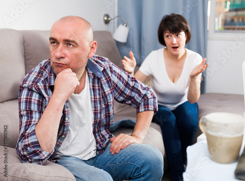 Portrait of upset mature man sitting, woman quarrelling on background at home © JackF