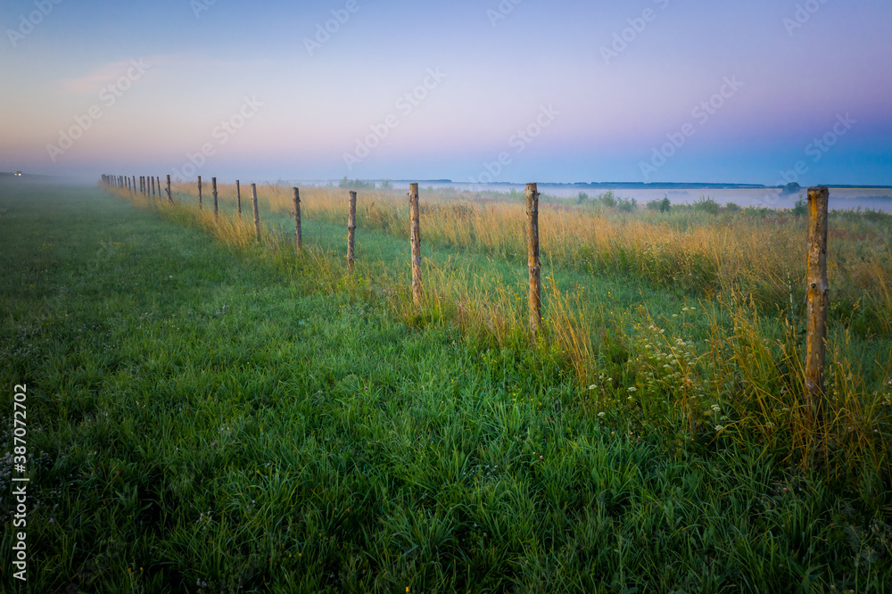 Morning fog in the meadows during sunrise in the countryside. Rural landscape with a fog on the geen field. the fence stretching into the distance.