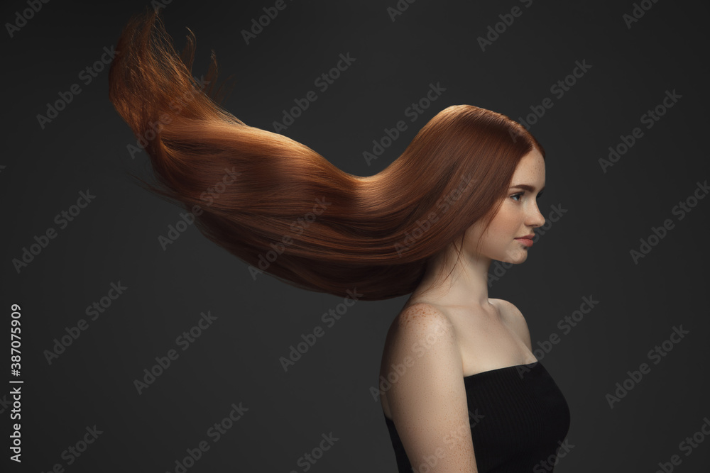 Beautiful model with long smooth, flying red hair isolated on dark grey studio background. Young caucasian girl with well-kept skin and hair blowing on air. Concept of salon care, beauty, fashion. Beautiful model with long smooth, flying red hair isolated on dark grey studio background. Young caucasian girl with well-kept skin and hair blowing on air. Concept of salon care, beauty, fashion.