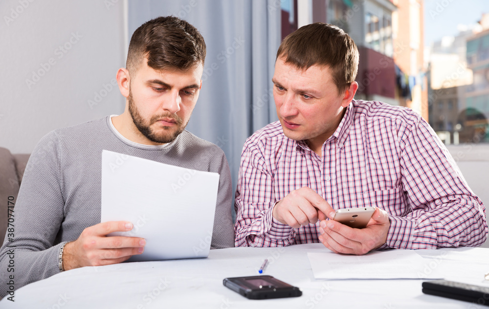 Two men in serious discussion analyzing documents together at home table, using phone