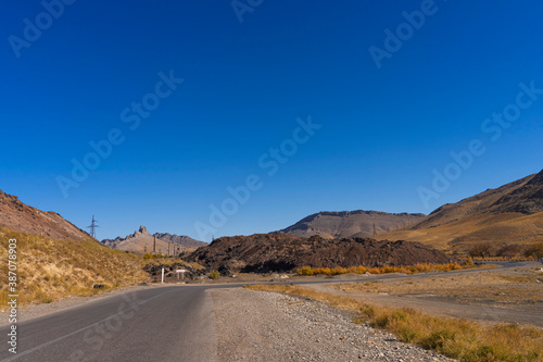 The road leads to the mountains. Mountain highway. Asphalt texture. Abandoned old factory in the background. Mountains and hills in autumn. Dry yellow grass. Blue sky. Mountain plants on the roadside 