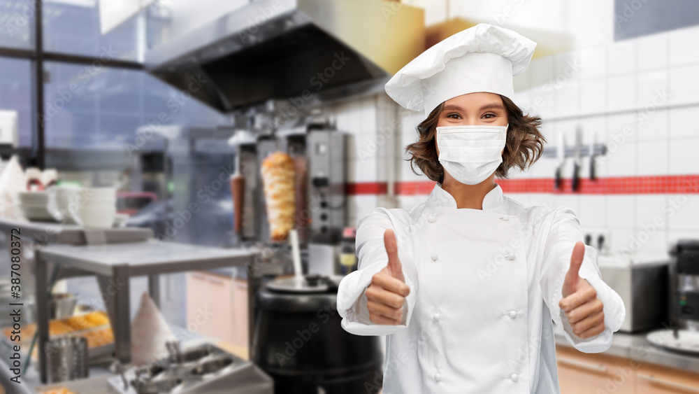 cooking, culinary and health concept - female chef in toque wearing face protective medical mask for protection from virus disease showing thumbs up over kebab shop kitchen background