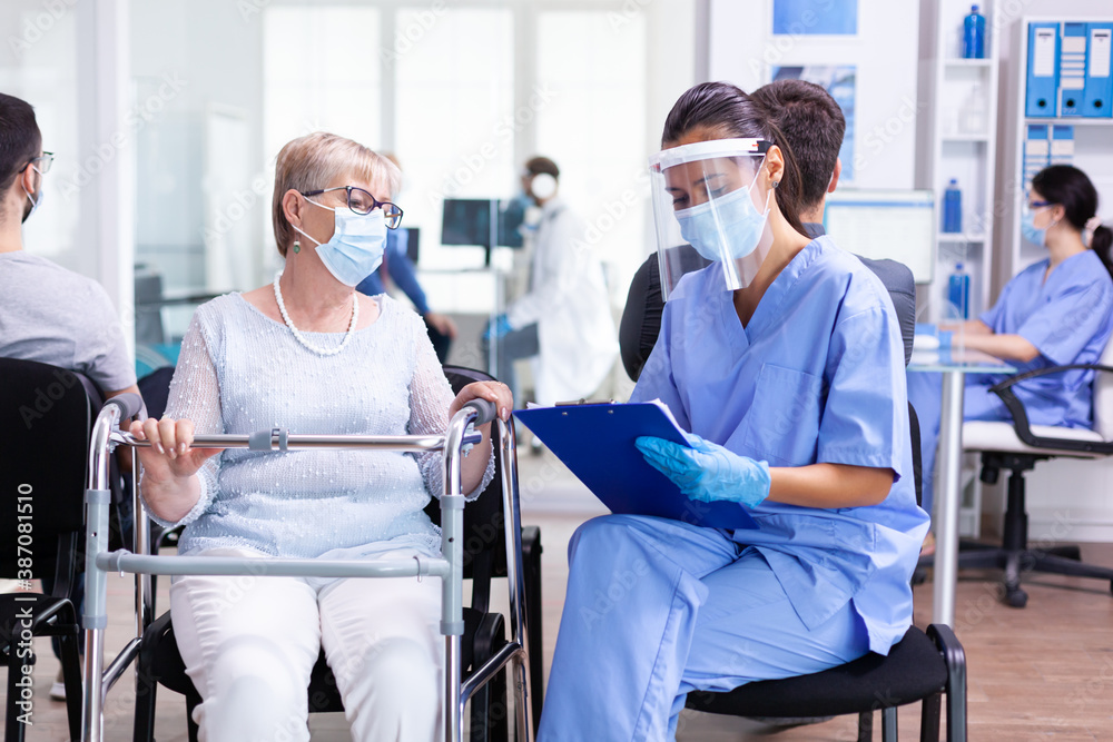 Disabled woman using walking frame in hospital waiting room wearing face mask against coronaviurs having a conversation with medical stuff. Patient and nurse in waiting area. Disabled woman using walking frame in hospital waiting room wearing face mask against coronaviurs having a conversation with medical stuff. Patient and nurse in waiting area.