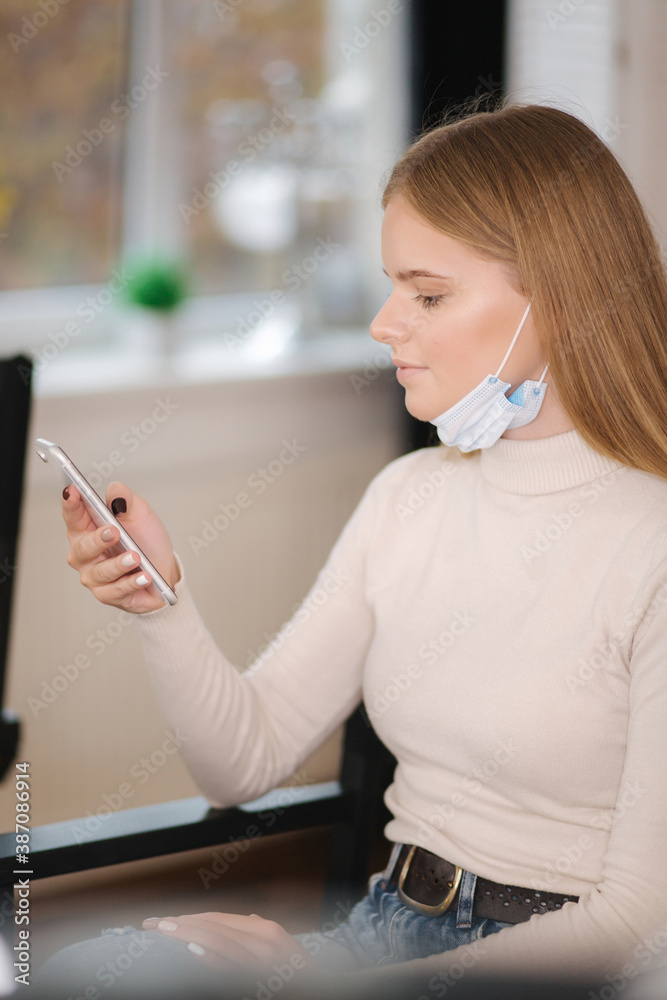 Female customer in beauty salon sitting on hight chair in medical mask on chin and use phone. Beautiful young blond hair woman wiating for brow master - obrazy, fototapety, plakaty 