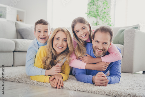 Photo of positive cheerful mommy daddy lying carpet hug two kids boy girl piggyback in house indoors
