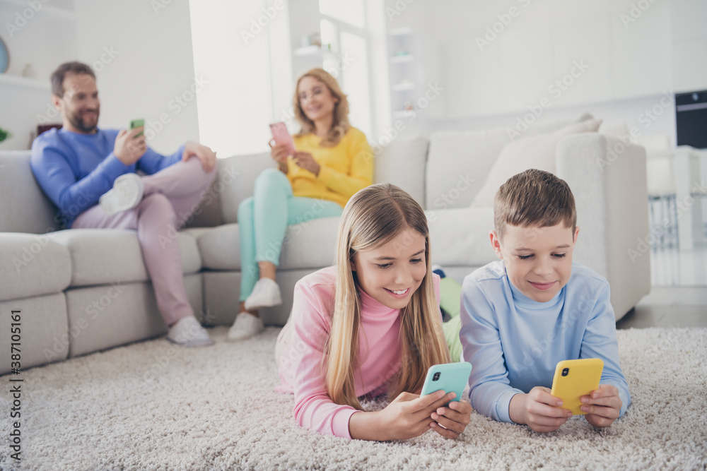 Full length photo of family people mommy daddy sit sofa two little kids lying carpet texting on smartphone in house indoors Full length photo of family people mommy daddy sit sofa two little kids lying carpet texting on smartphone in house indoors