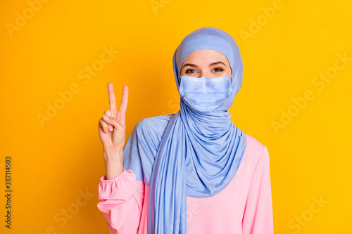 Close-up portrait of nice attractive cheerful muslimah lady showing v-sign wearing blue face mask isolated over bright yellow color background