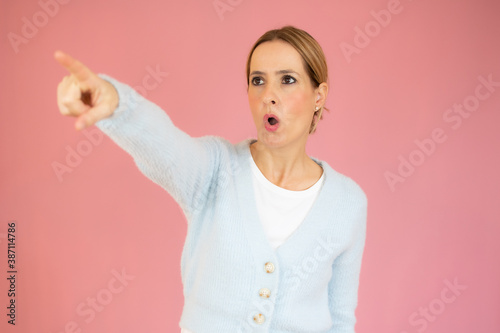Young beautiful woman wearing casual sweater over pink isolated background Pointing with finger surprised ahead, open mouth amazed expression, something on the front