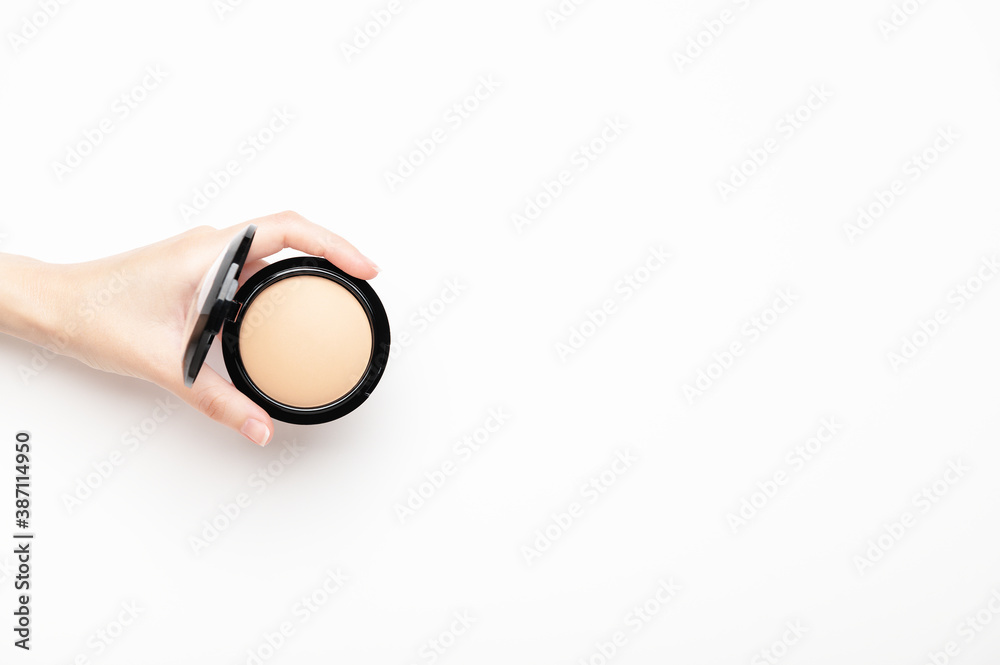 Obraz premium A female hand holds a compact beige powder on a white background, top view. Makeup nude product for perfect skin tone. Cosmetic, concealer, skin corrector. Copy space