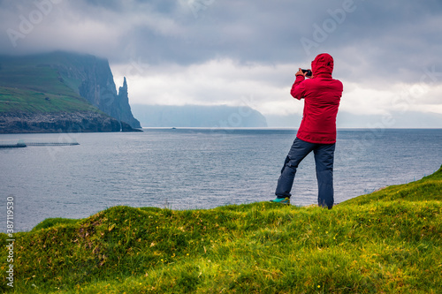 Tourist takes picture of Trollkonufingur cliffs on Vagar island. Dramatic summer scene of outskirts of Sandavagur village, Faroe Islands, Kingdom of Denmark, Europe.