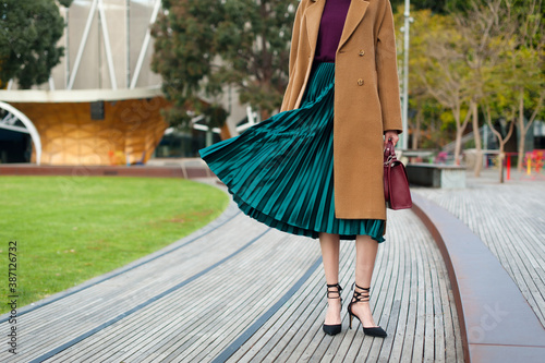 Fashionable young woman wearing green pleated midi skirt, sweater, high heel shoes, beige wool coat and holding burgundy handbag in hand on the city street. Street style.