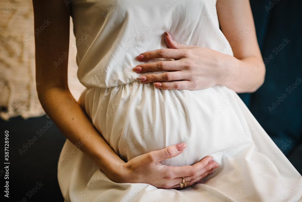 Pregnant woman in dress holds hands on belly at home. Maternity, pregnancy, motherhood, preparation and expectation concept. Beautiful tender photo of pregnancy indoors. Close up.