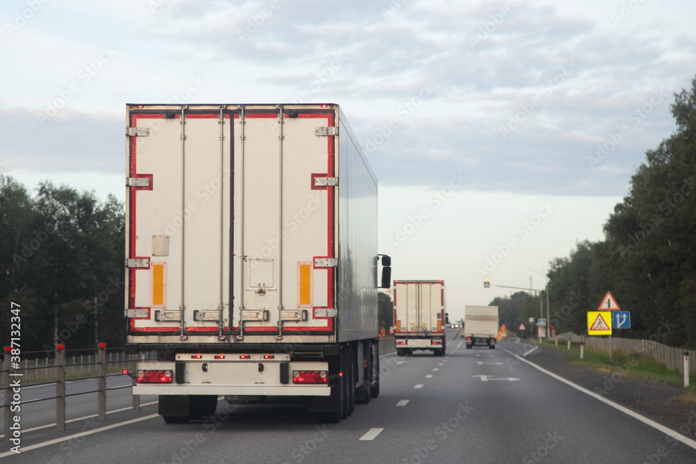 European heavy trucks with white semi trailers van drive on left side of two lane suburban asphalted highway road, back view at summer evening on forest and sky background, transportation logistics