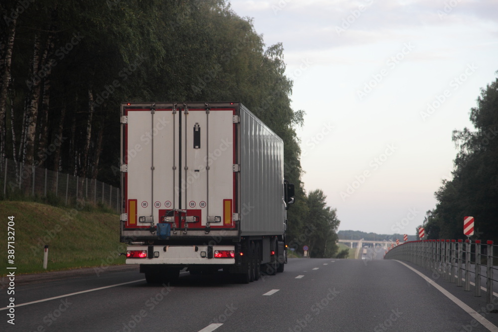 European heavy truck with white semi trailer van drive on left side of two lane suburban asphalted one way road, rear view at summer evening on forest and sky background, transportation logistics