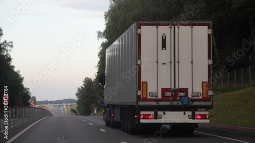 European heavy truck with white semi trailer van drive on two lane suburban asphalted highway road, back view at summer evening on forest and sky background, transportation logistics