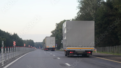 European trucks with gray awning van move on two lane suburban asphalted highway road, back view at summer evening on forest and sky background, cargo transportation logistics