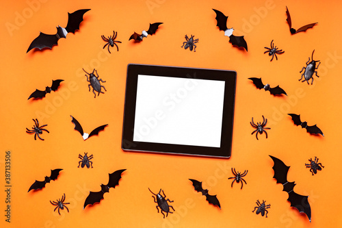 Halloween composition. Black bats, spiders, flies and bugs on orange background. Black empty tablet screen, mock up. Happy Halloween, trick or treat party concept. Flat lay, top view, copy space