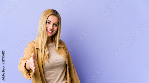 Young blonde caucasian woman stretching hand at camera in greeting gesture.