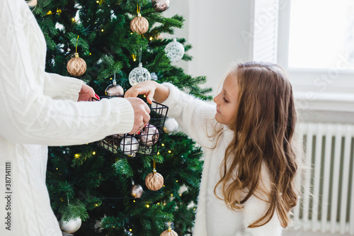 Mother and daughter stand near the Christmas tree, decorate the Christmas tree, light hair and white sweaters, beautiful decorations, holiday mood