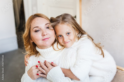 Mother and daughter, girl sitting on the sofa in light sweaters , hugs, emotions bright interior, winter atmosphere, on the eve of Christmas