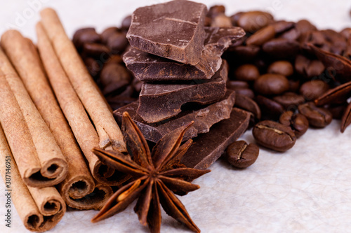 Stack of chocolate bar pieces with heap of roasted coffee beans and spices, cinnamon sticks and anise stars on a white table. Macro shot.