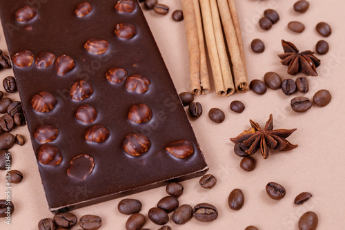 Chocolate bar with nuts on the beige background with coffee beans and spices. Roasted coffee beans, cinnamon sticks and anise stars. Chocolate bar with hazelnuts. 