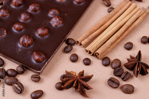 Chocolate bar with nuts on the beige background with coffee beans and spices. Roasted coffee beans, cinnamon sticks and anise stars. Chocolate bar with hazelnuts. 