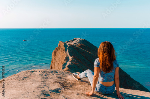 A young woman with lonh hair  sits on the slope of Cape chameleon with an amazing panoramic view of the hills and the sea from above © KseniaJoyg