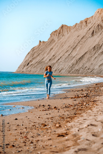 A young brunette woman in jeans runs along a picturesque sandy beach with a view of the Cape Chameleon rock in Crimea